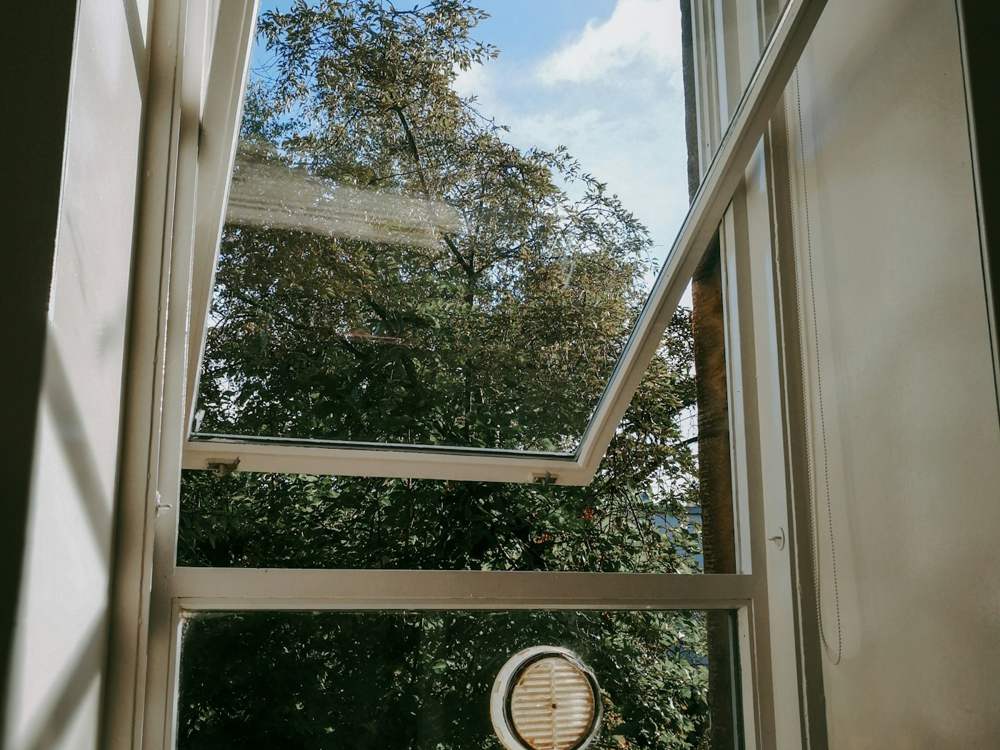 Fenêtre blanche ouverte en position basculée, vue de l’intérieur sur des arbres