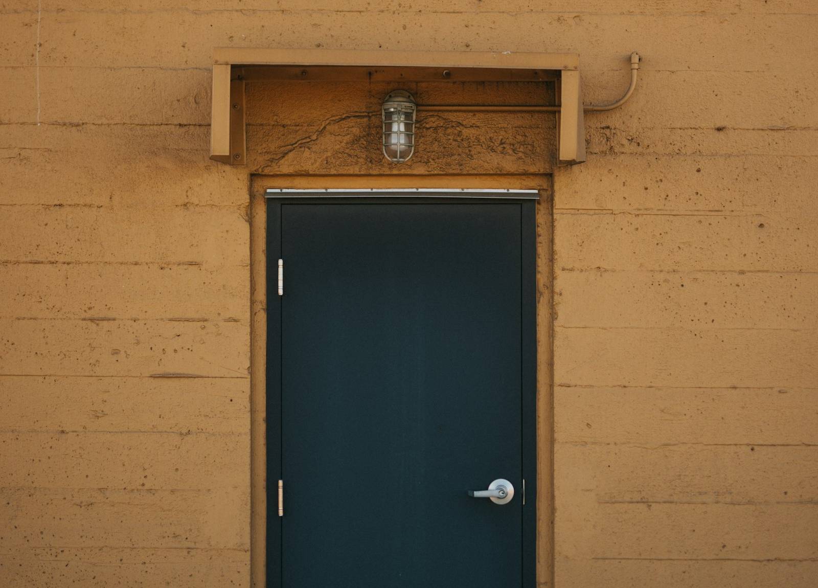 Porte de service pleine bleu foncé sur un mur ocre, sous une applique