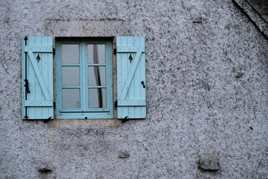Fenêtre aux volets battants bleus à barres et écharpe sur un mur crépi